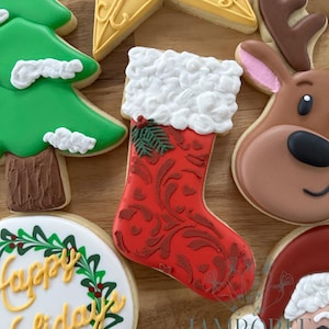 Op de afbeelding: Assortiment van versierde kerstkoekjes. Een rood sokkoekje met witte afwerking en een groene tak is omgeven door een groene kerstboom, een gele ster, een rendier en een koekje met de tekst "Happy Holidays".