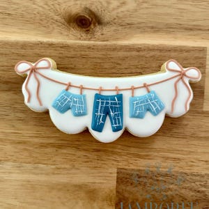 May include: A decorated sugar cookie shaped like a clothesline with baby clothes. The cookie is white with blue and white plaid shorts and trousers hanging from a string. The ends of the clothesline have pink bows.