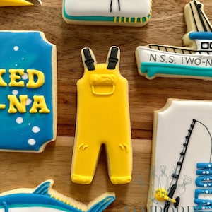 Puede incluir: Una colección de galletas de azúcar decoradas con temática de pesca. Las galletas incluyen un mono amarillo, un pez, un barco y una caña de pescar. Las galletas están decoradas con glaseado azul, amarillo, blanco y negro.