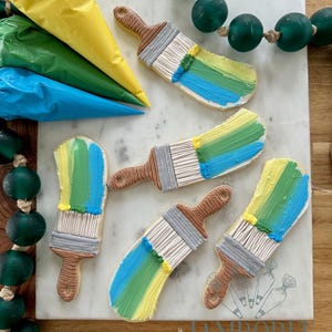 May include: Decorated sugar cookies shaped like paintbrushes with colourful frosting strokes in yellow, green, and blue. The cookies are arranged on a white marble surface with icing bags and green bead garland.
