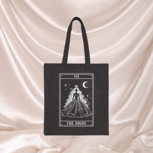 May include: Black tote bag featuring a white tarot card design. The card depicts a bride with a crown, long flowing hair, and a dress, with a crescent moon and stars. The text "VII" and "THE BRIDE" are also visible.