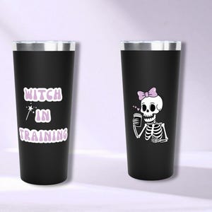 Può includere: Due bicchieri termici neri. Uno presenta la scritta "Witch in Training" in lavanda, con una bacchetta magica. L'altro ha uno scheletro con un fiocco rosa, che tiene una tazza di caffè.