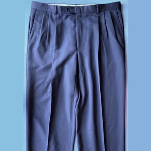 May include: Navy blue pleated trousers with a button closure and side pockets. The pants have a cuffed hem. The fabric appears to be a smooth, woven material. The trousers are displayed against a light blue background.