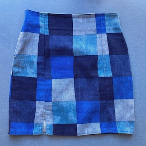 May include: A denim mini skirt with a patchwork design in shades of blue and gray. The skirt features a high waist and a small slit at the front hem. The fabric has a textured appearance.