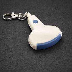 May include: A white and blue keychain shaped like an ultrasound probe. The keychain has a silver clasp and a small blue button on top. The bottom edge is blue. The background is black.