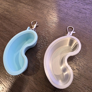 May include: Two kidney-shaped keychains with silver clasps. One is light blue, the other is translucent white. Both have a textured interior and a smooth, rounded exterior. The keychains are likely 3D-printed.