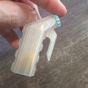 May include: A 3D-printed keychain in the shape of a small spray bottle. The keychain is white with a light blue cap and a handle. The keychain is held by a person's hand.