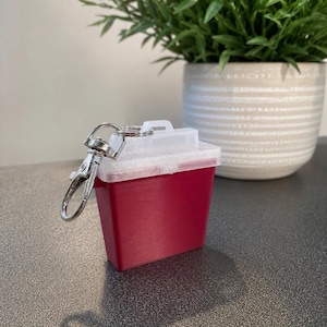 May include: A small, rectangular, deep red container with a translucent white lid and a silver-toned metal lobster clasp keychain attached to the top. The object features a rigid, box-like structure with a flat base and a secure, hinged-style closure.