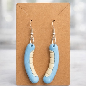 May include: A pair of blue and cream-colored earrings shaped like a curved, segmented design. The earrings hang from silver-colored hooks and are displayed on a brown cardboard backing.