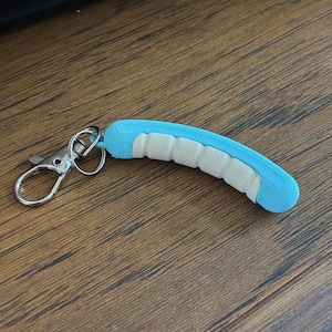 May include: A blue and white worm-shaped keychain with a silver clasp and key ring. The keychain is made of a light blue material with a white segmented design. The keychain is on a wooden surface.