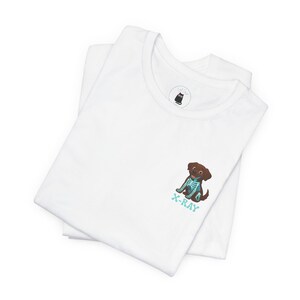 May include: White t-shirt featuring a cartoon brown dog with a turquoise X-ray design. The text "X-RAY" is below the dog. The shirt is folded, showing the collar and a small logo.