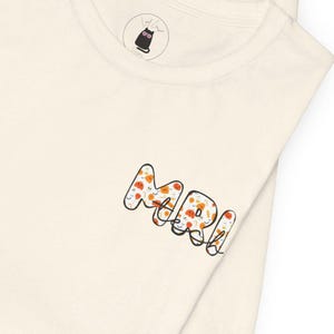 Pode incluir: Camiseta creme com a palavra "MARI" em letras de bolha laranja e brancas, com um padrão de rostos sorridentes. Um pequeno logotipo de gato preto é visível perto da gola.