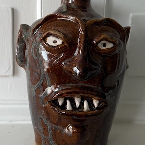 May include: A brown ceramic jug with a face sculpted on the front. The jug has a wide mouth with white teeth, white eyes, and small ears. The jug is sitting on a white shelf.