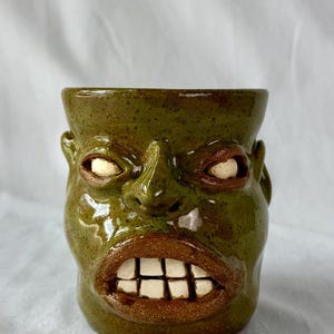 May include: A ceramic mug shaped like a human face with a glossy olive green glaze. The mug features prominent facial features including a nose, ears, and a wide mouth with visible white square teeth and brown lips, plus white eyes.
