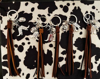 May include: Six keychains with silver charms and brown leather tassels. Each keychain features a silver ring, a leather tassel, and multiple charms, including cowboy boots, hats, and horseshoes. The keychains are arranged on a black and white cowhide pattern.