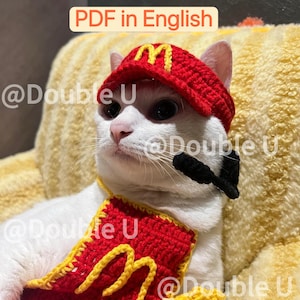 Puede incluir: Un gato blanco con un gorro y babero de ganchillo rojos y amarillos, que se asemejan a un uniforme de comida rápida. El gorro y el babero presentan la letra "M" en amarillo. El texto "PDF in English" está en la parte superior de la imagen.