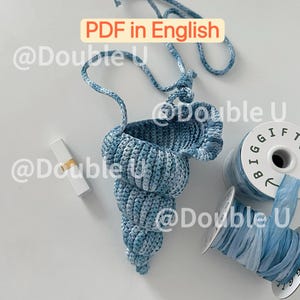 Op de afbeelding: Een handgemaakte, blauw-witte gehaakte schelptas met een lange band. Naast de tas een lippenstift en spoelen met blauw lint. De tekst "PDF in English" is zichtbaar.