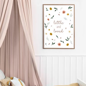 Könnte beinhalten: Ein gerahmter Druck mit dem Schriftzug "little and loved" in zarter Schrift, umgeben von floralen und Blattillustrationen in Rosa, Gelb und Grün. Der Druck hängt an einer weißen Wand, mit einem zartrosa Vorhang und Kissen im Vordergrund.