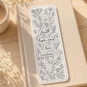 Könnte beinhalten: Schwarz-weiß Lesezeichen mit floralen Illustrationen und den Worten "Glaube, Hoffnung und Liebe" und "1. Korinther 13,13". Das Lesezeichen liegt auf einem beigen Buch.