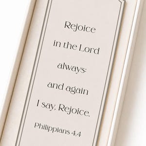Könnte beinhalten: Ein rechteckiges Lesezeichen mit cremefarbenem Hintergrund und grauem Rand. Der Text lautet: Rejoice in the Lord always: and again I say, Rejoice. Philippians 4,4. Unten ist das Wort Segenswünsche mit einem kleinen kreisförmigen Symbol abgedruckt.