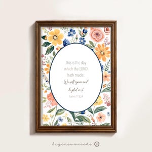 Könnte beinhalten: Ein gerahmter Blumendruck mit einem Aquarelldesign. Der Druck zeigt eine Vielzahl farbenfroher Blumen in Gelb-, Blau- und Pfirsichtönen. Der Text innerhalb des ovalen Rahmens lautet: "This is the day which the LORD hath made; We will rejoice and be glad in it Psalms 118,24."