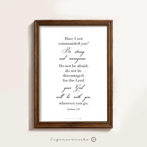 Könnte beinhalten: Ein gerahmter Druck mit einem dunkelbraunen Holzrahmen. Der Druck zeigt eine elegante Schrift mit dem Text "Have I not commanded you? Be strong and courageous..." und dem Bibelvers Josua 1,9. In der unteren rechten Ecke steht der Text "Segenswünsche".