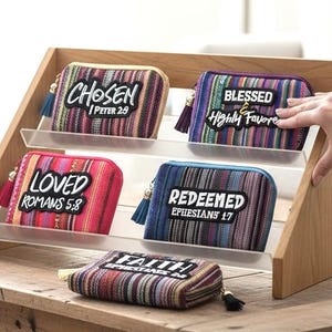 May include: A wooden display stand holds several colorful zippered pouches with inspirational text. The pouches feature striped patterns in various colors and include phrases like "Chosen," "Blessed," "Loved," "Redeemed," and "Faith."