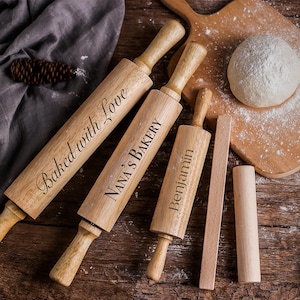 May include: Wooden rolling pins with engraved text, including "Baked with Love," "Nana's Bakery," and "Benjamin." A small rolling pin, a dough scraper, and a dough roller are also present. The items are on a wooden surface.