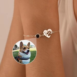 May include: A silver bracelet featuring a heart-shaped charm with a paw print and the name "Buddy". The bracelet also includes a circular black stone and a chain. A circular image of a dog is in the background.