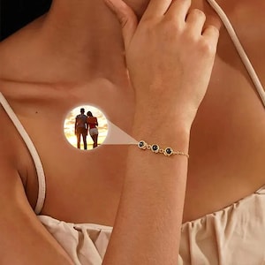 May include: A gold bracelet with four black circular stones. The bracelet is on a person's wrist. A small circular image of a couple is in the background.