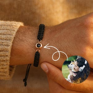 May include: Black braided bracelet with a gold-coloured accent and a black stone. The bracelet is on a wrist. A small circular image shows a person with a dog.