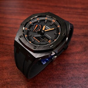 May include: A black G-Shock watch with orange accents on a dark wooden surface. The watch features a digital display and a black rubber strap. The watch face has a textured design and visible screws.