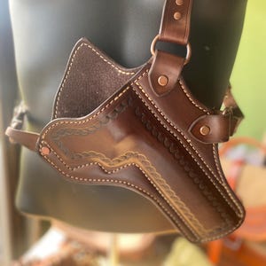 Puede incluir: Una funda de hombro de cuero marrón con costuras decorativas y un patrón floral. La funda tiene correas ajustables y remaches de cobre. El diseño es visible en un maniquí negro.