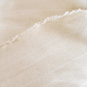 May include: Close-up of a piece of off-white fabric with a woven texture. The fabric has a raw edge.