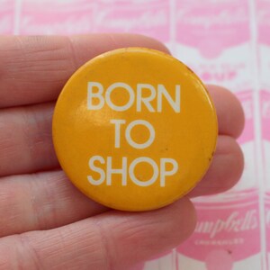 May include: A yellow button with the text "BORN TO SHOP" in white letters.