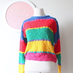 May include: A colorful, striped sweater in shades of red, blue, green, yellow, and pink. The vintage knitwear is displayed on a mannequin. The sweater has long sleeves and a round neckline. The background includes a white wall and a door.