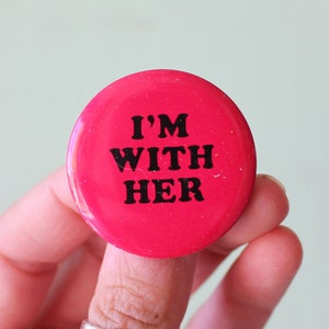 May include: A red button with the text "I'M WITH HER" in black lettering.