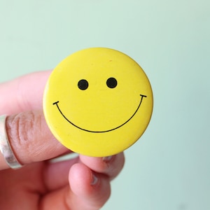 May include: A yellow smiley face pin with a black outline. The pin is being held by a hand.