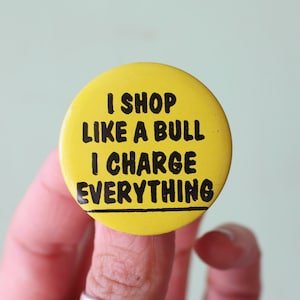 May include: Yellow button with black text that reads "I shop like a bull I charge everything".