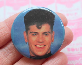 May include: A round, blue button featuring a smiling man with dark hair and hoop earrings. The button has a retro aesthetic, likely from the 1980s or 1990s. The background is a pink pattern with Campbell's Soup cans.