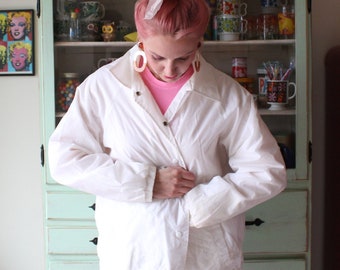 May include: A woman with pink hair is wearing a white windbreaker jacket with a pink shirt underneath. She is standing in front of a light green cabinet with shelves filled with various items. There is a red refrigerator with the word "GALANZ" on the front.