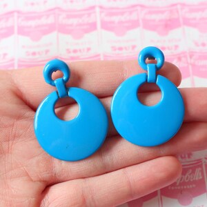 Puede incluir: Un par de pendientes colgantes azules con un gran diseño circular. Los pendientes tienen un estilo simple y moderno.