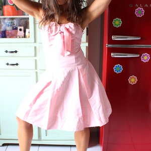 May include: A woman in a pink dress with a bow at the neckline stands in a kitchen with a red refrigerator with the word "GALANZ" on it. The refrigerator has colorful flower stickers on it.