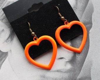 Peut inclure: Une paire de boucles d'oreilles en forme de cœur orange avec des crochets dorés. Les boucles d'oreilles sont présentées sur une carte noire.