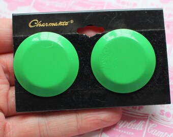 Op de afbeelding: Een paar groene plastic oorbellen op een zwarte kaart met het woord "Charmante" erop gedrukt.