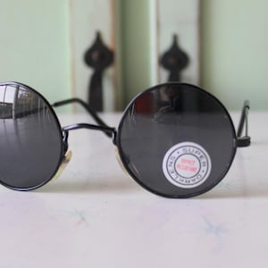 May include: Black round sunglasses with a black frame and dark lenses. The sunglasses have a small sticker on the right lens that says "100% UV Protection".