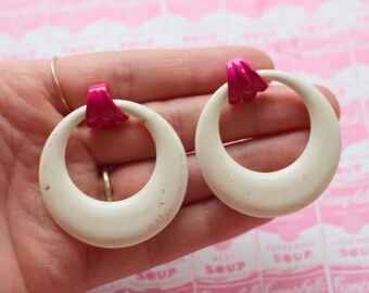 May include: A pair of white hoop earrings with a pink accent. The earrings are large and have a rounded shape.