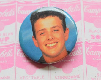 May include: A blue and white button featuring a portrait of a young man with brown hair. The button is on a background of pink and white Campbell's Soup cans.