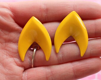 May include: A pair of yellow enamel earrings shaped like a flame. The earrings are in the shape of a pointed oval with a rounded top and a pointed bottom.