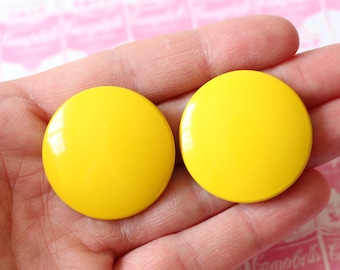 May include: Two bright yellow, round earrings are held in a hand. The earrings are glossy and appear to be made of plastic or resin. The background is a pink and white pattern with text that reads "Campbell's Soup".